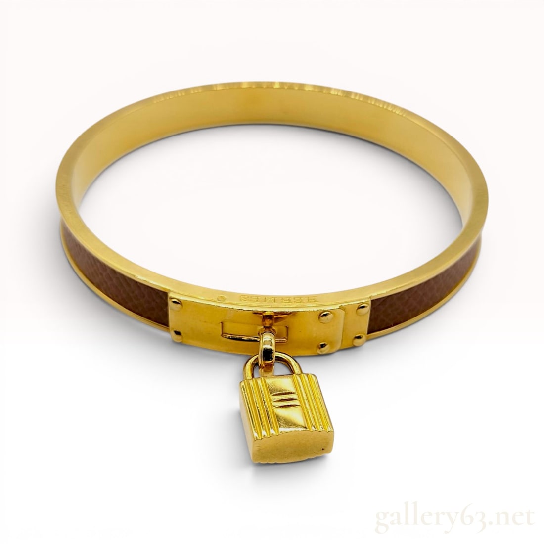 Hermes Gold-Tone and Brown Leather Kelly Cadena Lock Bangle Bracelet: A Hermes Kelly Cadena lock bangle bracelet featuring a gold-tone metal frame with a textured brown leather inlay. The design is modeled after the signature turn-lock hardware of the Hermes Kelly bag, 