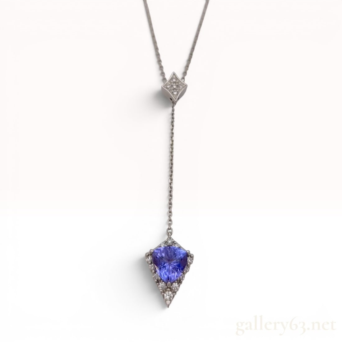 Platinum 950 Trillion-Cut Tanzanite and Diamond Lariat Pendant Necklace with Box: This platinum lariat-style necklace features a trillion-cut blue-violet tanzanite as its primary stone. The tanzanite is secured in a three-prong setting and surrounded by a chevron-shaped halo accent