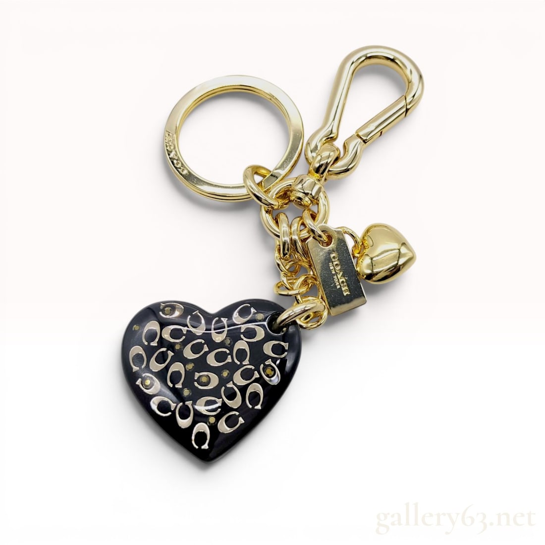 Coach Signature C Heart Bag Charm Keychain: This gold-tone metal accessory functions as both a keychain and a bag charm, featuring a primary heart-shaped pendant crafted from black resin. The resin heart is decorated with an inset silver-tone C