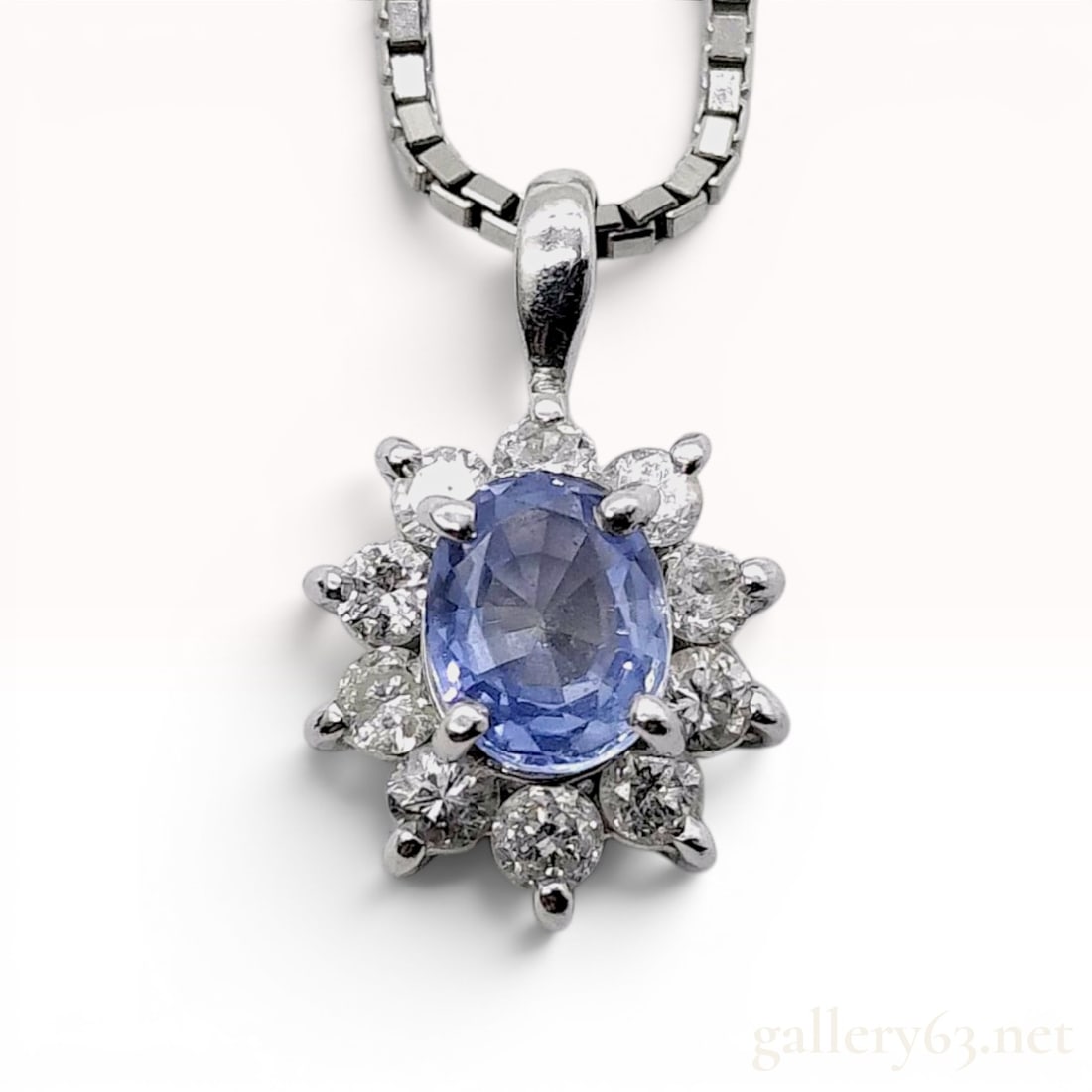 Platinum Tanzanite and Diamond Cluster Pendant Necklace: This platinum pendant necklace features a central oval-cut light blue tanzanite set within a halo of eight round brilliant-cut diamonds. The pendant is suspended from a platinum box-link chain. The re