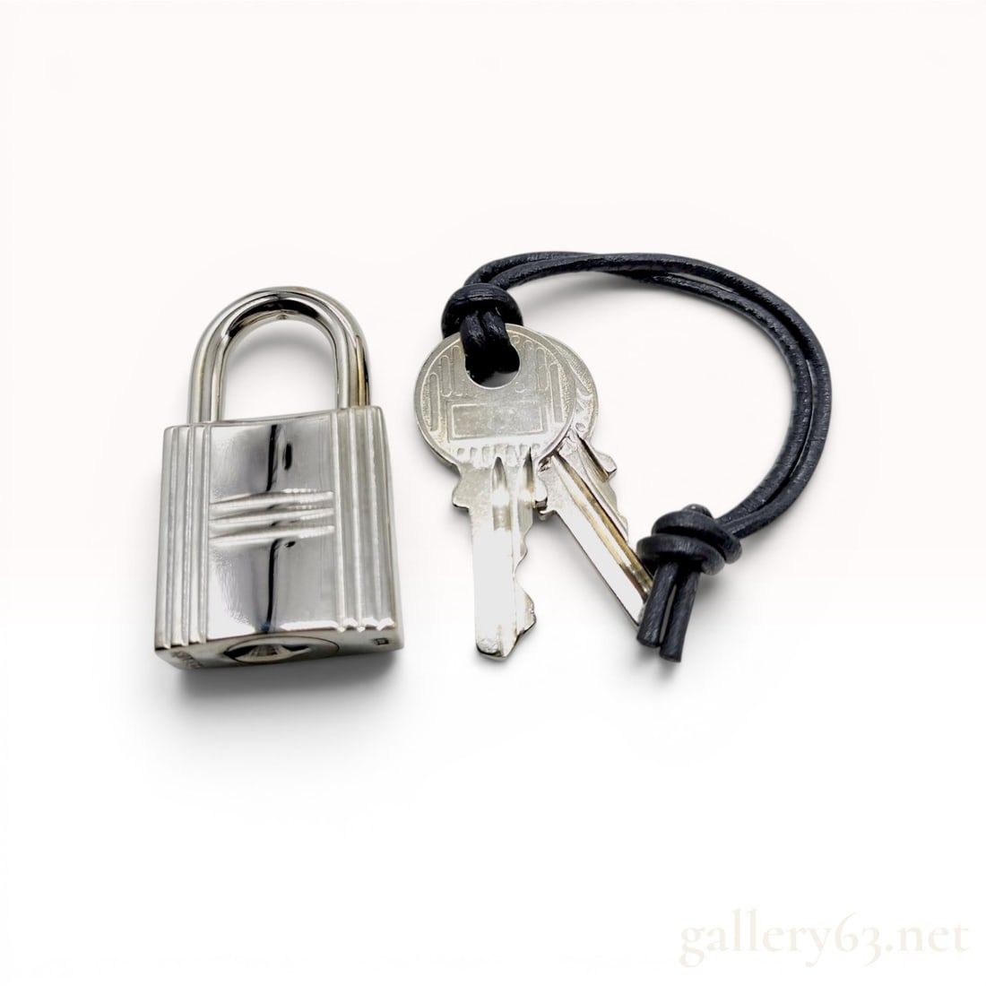 Hermes Palladium-Tone Padlock and Two Keys with Leather Cord: A silver-tone metal padlock and matching key set by Hermes, commonly referred to as a cadena. The rectangular lock features a polished finish and a debossed stylized H motif on the front. The base of 