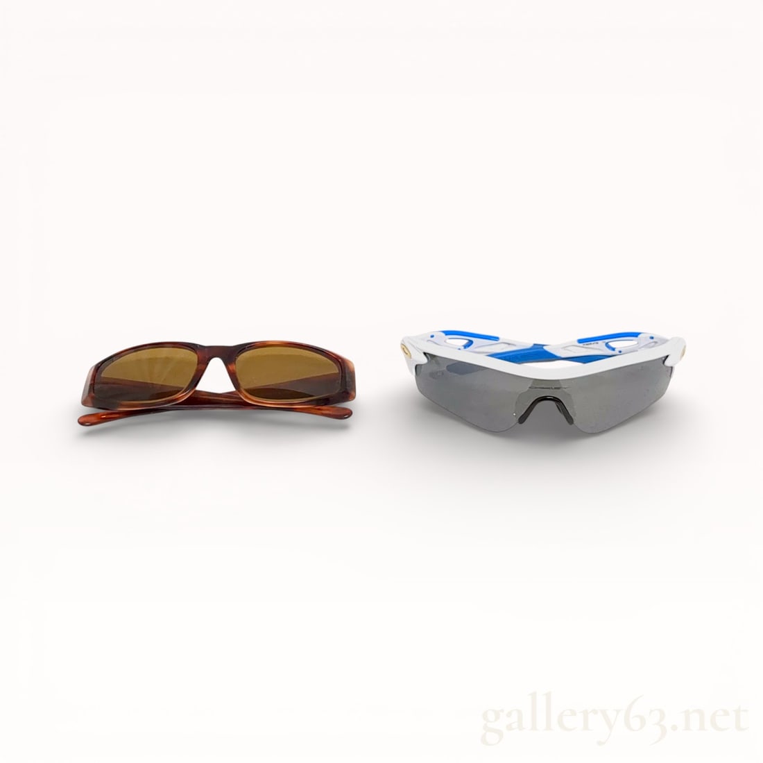Two Pairs of Designer Sunglasses by Ray-Ban and Oakley: A pair of Ray-Ban tortoiseshell acetate sunglasses and a pair of Oakley performance sport sunglasses. The Ray-Ban frames feature brown tinted lenses and a gold-tone swan emblem on the left temple, whi