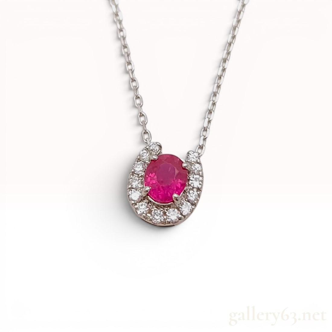 Ruby and Diamond Horseshoe Pendant Necklace in Platinum: Platinum pendant necklace featuring a central prong-set oval-cut ruby surrounded by a horseshoe-shaped halo of twelve round-cut diamonds. The pendant is suspended from a fine cable link chain with a s