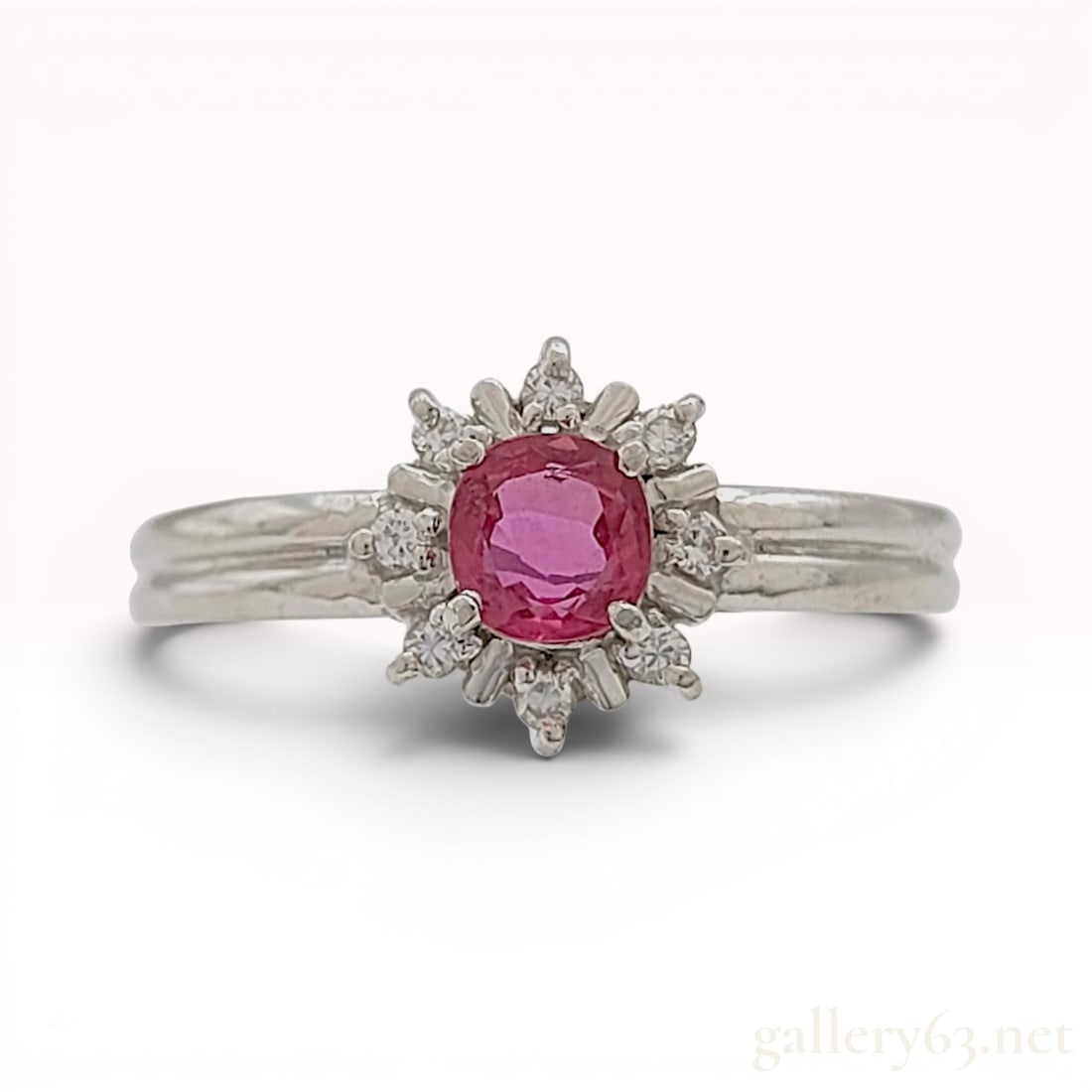 Platinum Pinkish-Red Ruby and Diamond Cluster Ring: A platinum cocktail ring featuring a sunburst-style cluster head. The center of the ring holds a faceted round-cut pinkish-red ruby secured in a four-prong setting. The interior of the band features a