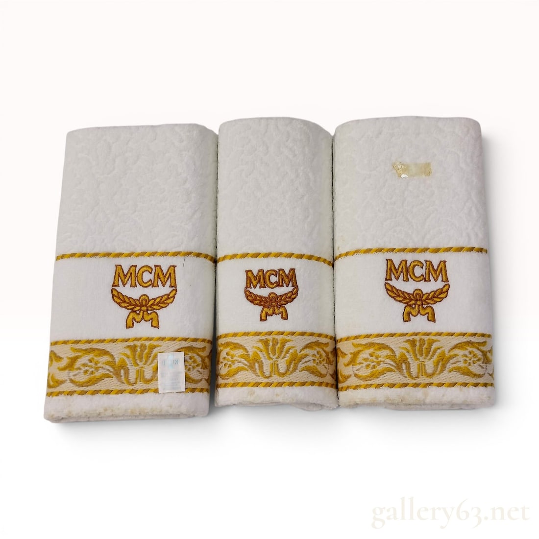A Trio of MCM Branded Hand Towels: A trio of white cotton hand towels featuring the MCM logo. Each towel is constructed from a white terry cloth fabric with a textured damask-style weave. The decorative elements consist of two woven go