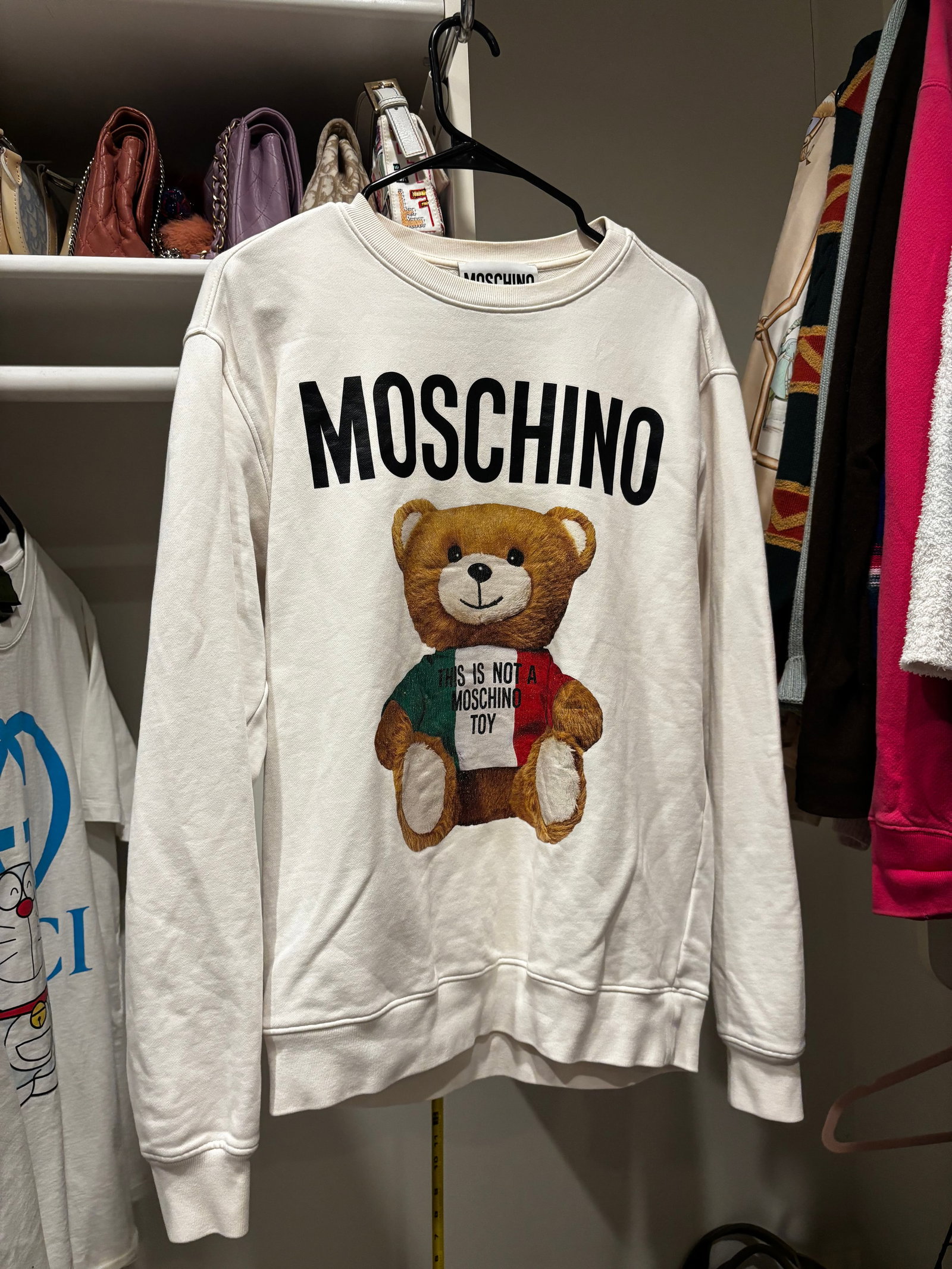 Moschino Couture Italian Flag Teddy Bear Crewneck Sweatshirt: A Moschino Couture long-sleeved crewneck sweatshirt in an off-white cotton jersey. The front displays a vertical black block-letter Moschino logo alongside the brand signature teddy bear mascot. The b