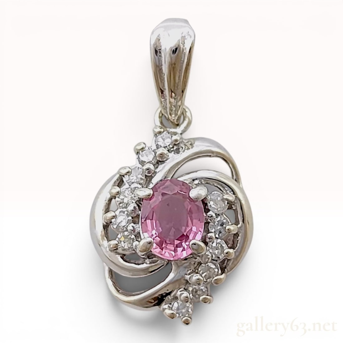14 Karat White Gold Pink Sapphire and Diamond Pendant: This jewelry pendant is crafted in 14 karat white gold and features a central oval-cut pink sapphire. The primary gemstone is held in a four-prong setting and is surrounded by a swirled bypass-style c