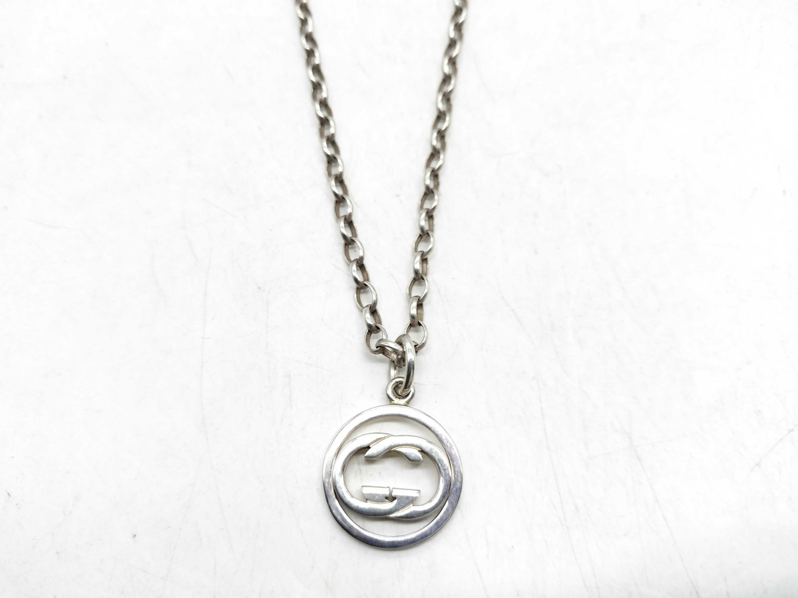 Gucci Sterling Silver Interlocking GG Logo Pendant Necklace: This is a sterling silver pendant necklace manufactured by Gucci. The piece features a rolo style chain composed of round links. Attached to the chain is a circular pendant containing a cut-out interl