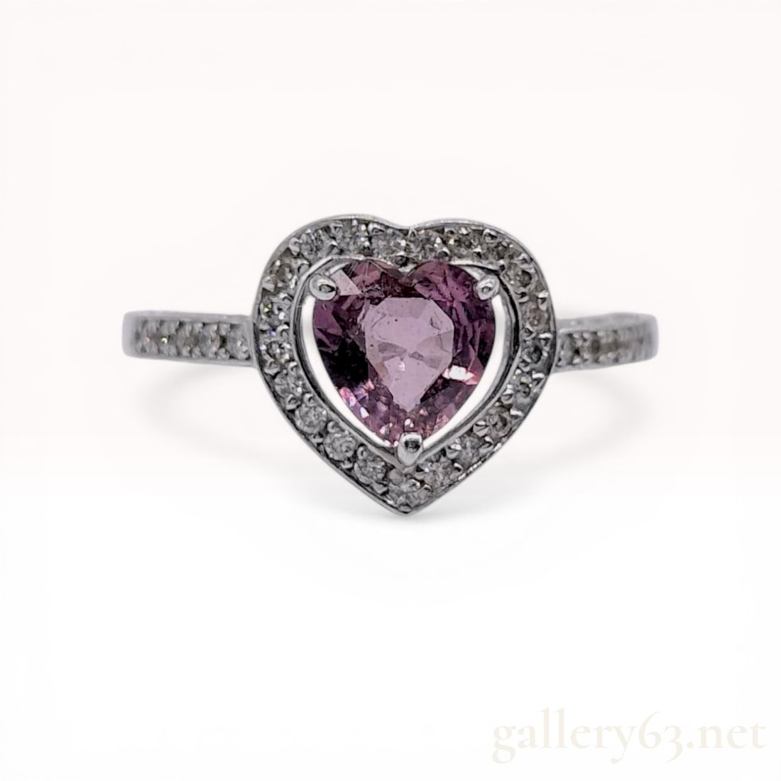 18 Karat White Gold Heart Shaped Sapphire and Diamond Halo Ring: 18 karat white gold ring featuring a faceted heart-shaped pinkish-purple sapphire within a diamond halo. The center stone is secured by three prongs, while round brilliant-cut diamonds are bead-set in