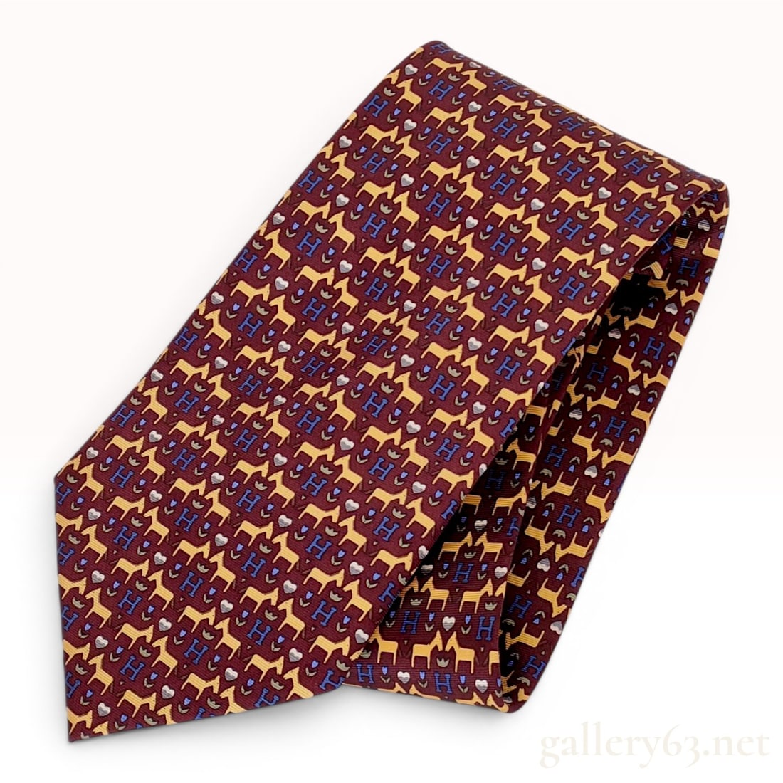 HERMES 100% Silk Burgundy Horse and H Logo Pattern Necktie: An Hermes 100% silk necktie featuring a burgundy ground with a whimsical repeating pattern of yellow stylized horses, blue H initials, light gray hearts, and small blue floral sprigs. The tie is const