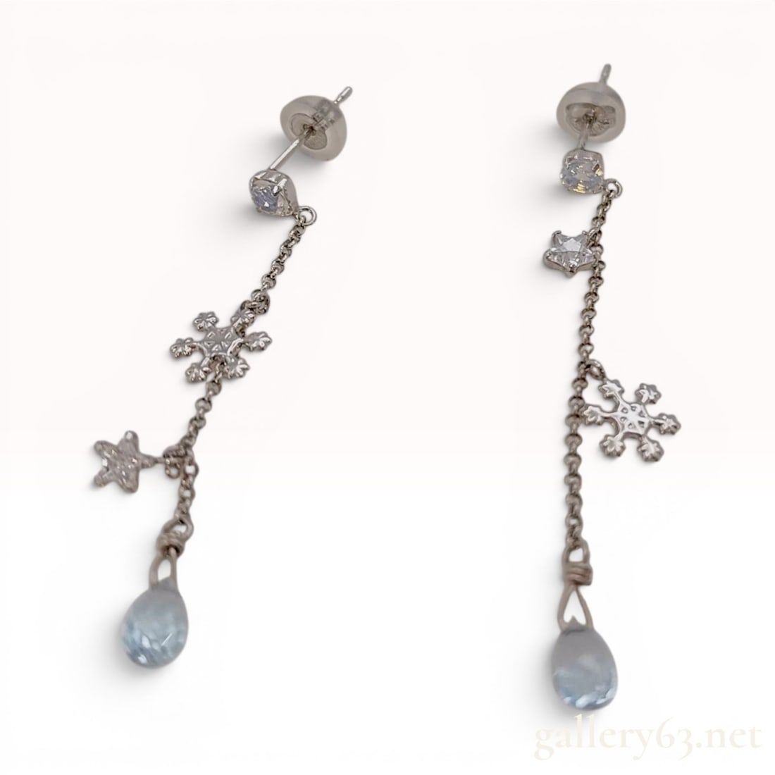 A Pair of 10K White Gold Celestial and Snowflake Drop Earrings: Each earring is constructed from 10 karat white gold and features a linear drop design with multiple charms. The arrangement begins with a round-cut clear stone at the stud, followed by a star-cut cle