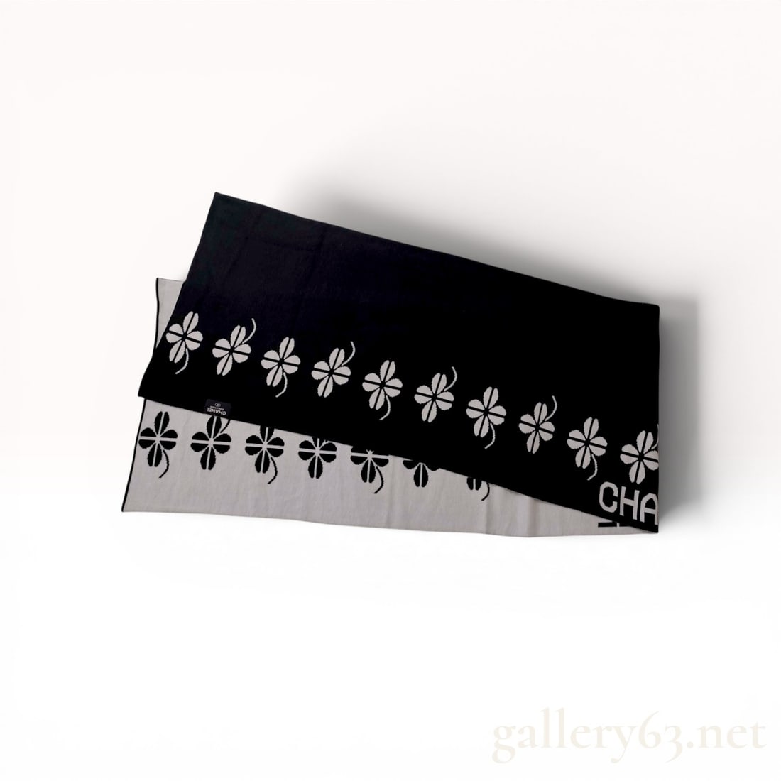 Chanel Boutique Reversible Black and White Knit Clover Motif Scarf: Chanel reversible knit scarf featuring a black and white graphic design. The textile displays a repetitive four-leaf clover motif alongside integrated bold Chanel typography on both sides. A black fab