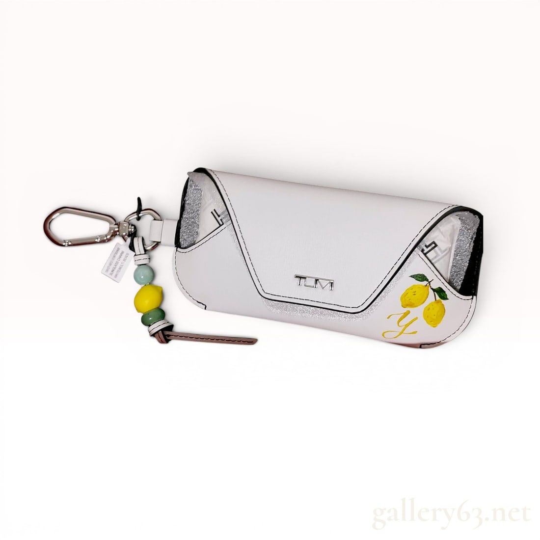 Tumi Belden White Leather Sunglass Case Charm with Lemon Motif: A Tumi Belden sunglass case and bag charm crafted from white textured leather with black glazed edges. The exterior is decorated with a printed yellow lemon and green leaf design and a yellow script l