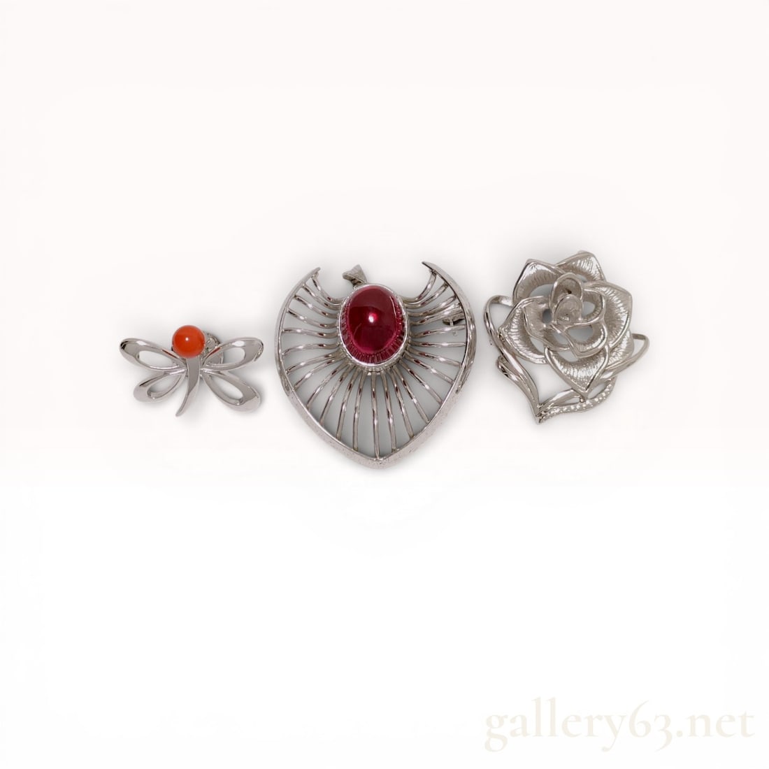 Three Silver Brooches and Pins Including Heart and Rose Motifs: A trio of silver jewelry pieces including two brooches and one lapel pin. The collection features a heart-shaped sunburst brooch with a ruby-red glass cabochon, a detailed rose blossom brooch, and a s