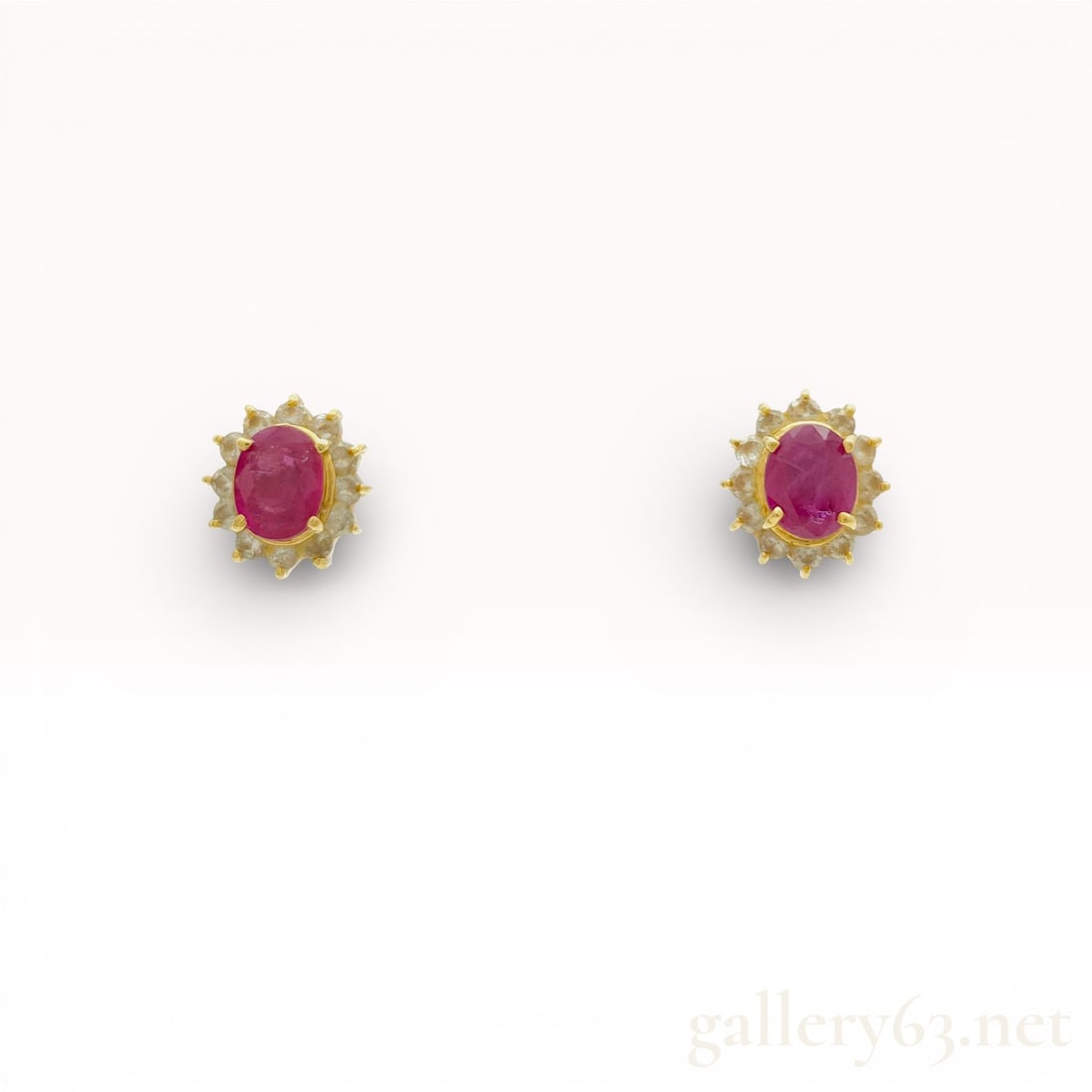 A Pair of 18K Yellow Gold Ruby and Diamond Halo Stud Earrings: A matched pair of ruby and diamond halo stud earrings set in 18 karat yellow gold. Each earring features a central prong-set oval-cut pinkish-red ruby surrounded by a sunburst frame of twelve round-cu