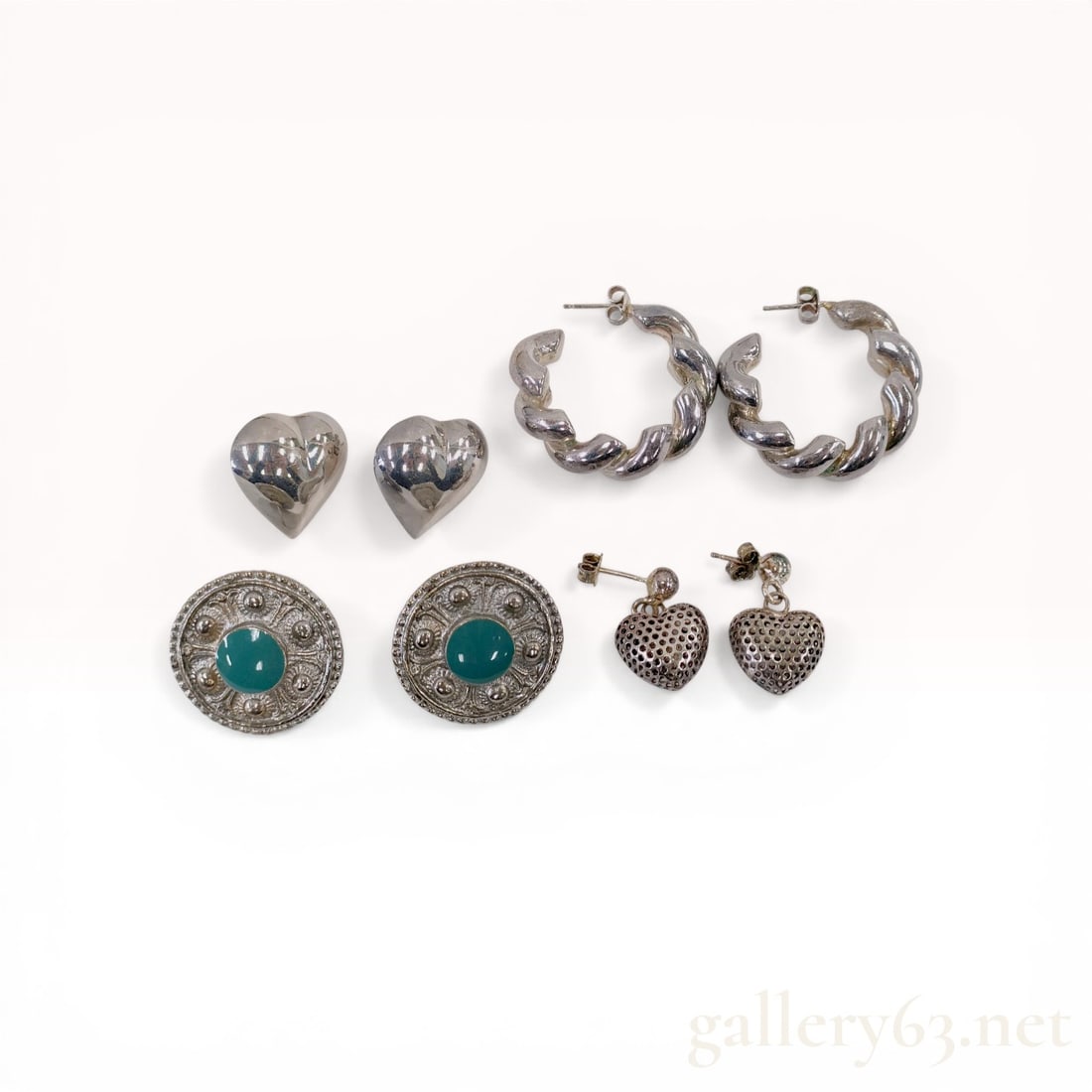 An Assortment of Four Pairs of Sterling Silver Earrings: An assortment of four matched pairs of sterling silver and silver-tone earrings. The collection includes puffed heart studs, heavy twisted open hoops, ornate button studs with green cabochons, and per