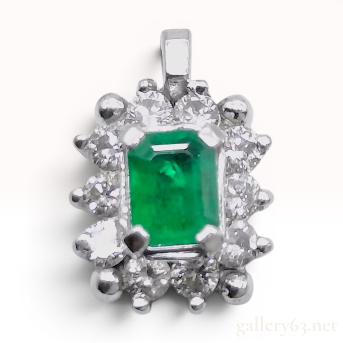 Platinum Emerald and Diamond Cluster Pendant: A platinum cluster pendant featuring a central rectangular emerald-cut green emerald. The center gemstone is secured by a four-prong setting and is bordered by a halo of ten round brilliant-cut colorl