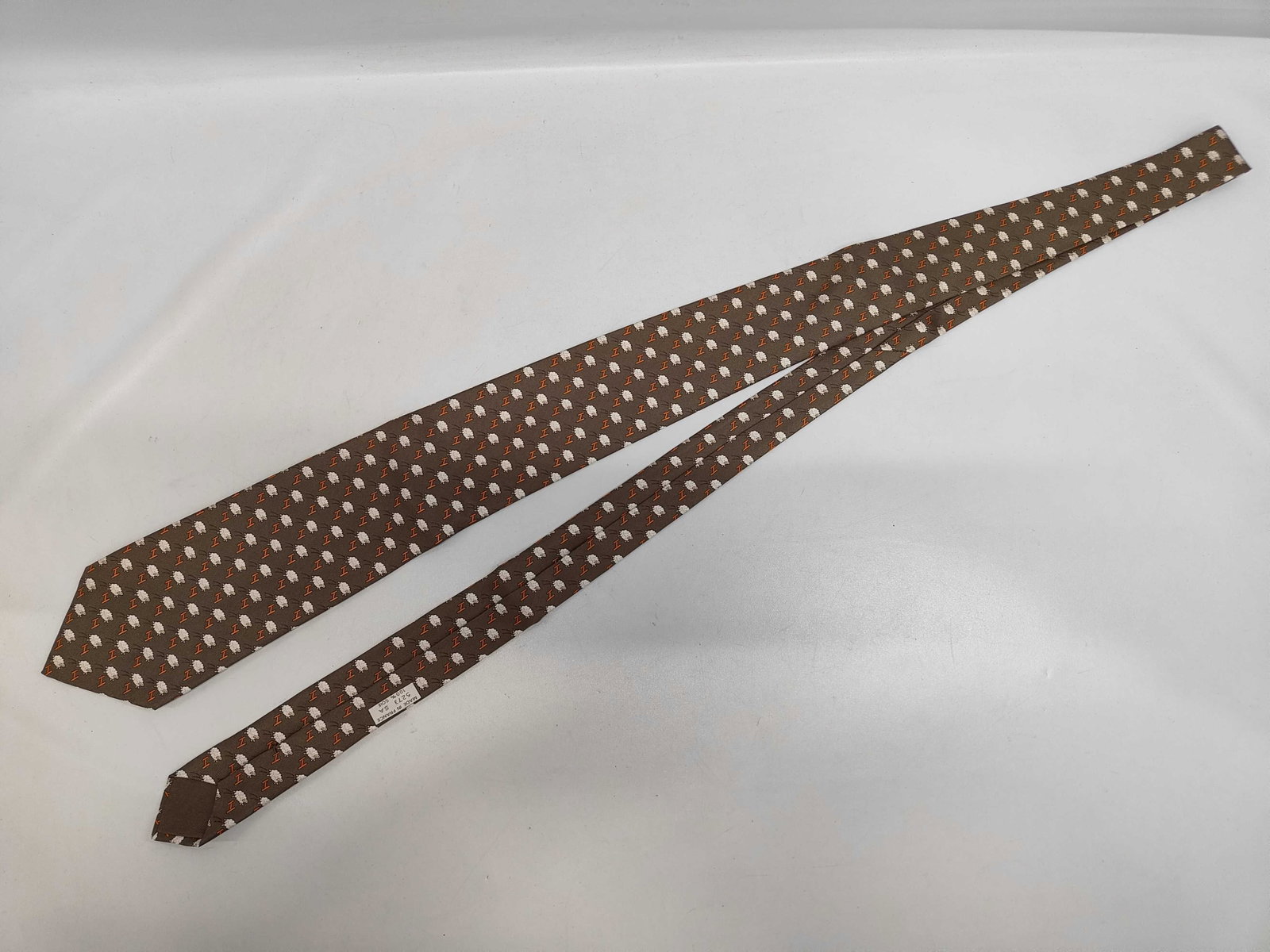 Hermes Taupe Silk Necktie with Sheep and H Logo Pattern: This genuine Hermes silk necktie features a whimsical repeating pattern of stylized white sheep and orange H logos set against a taupe background. The tie is constructed with a traditional three-fold 