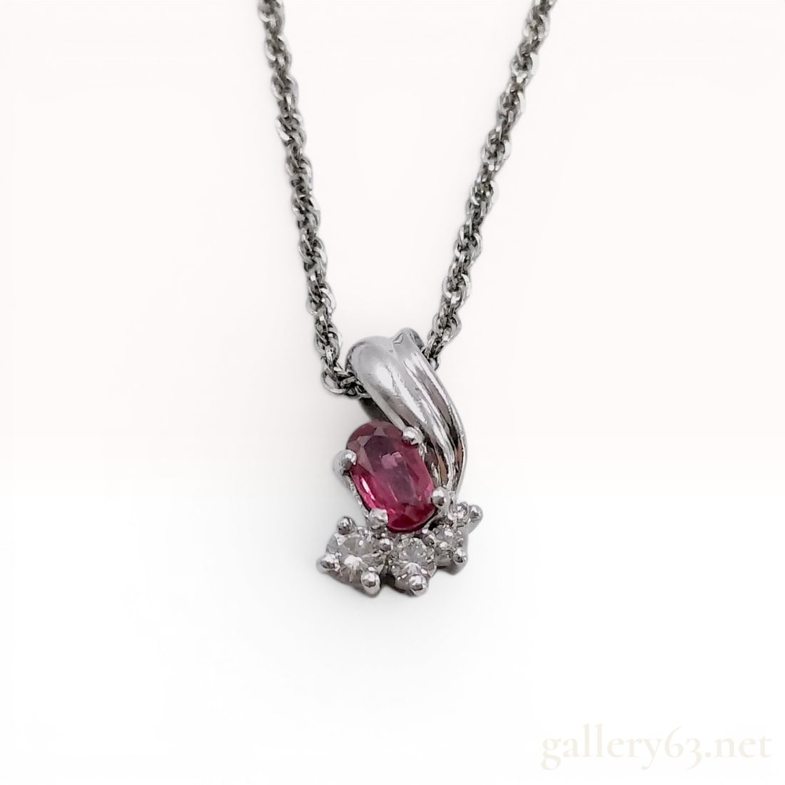 Platinum Necklace with Ruby and Diamond Pendant: A platinum necklace featuring a ruby-set pendant. The pendant consists of an asymmetrical, swirling platinum mount hallmarked Pt900. It is set with a central oval-cut faceted pinkish-red ruby with a w