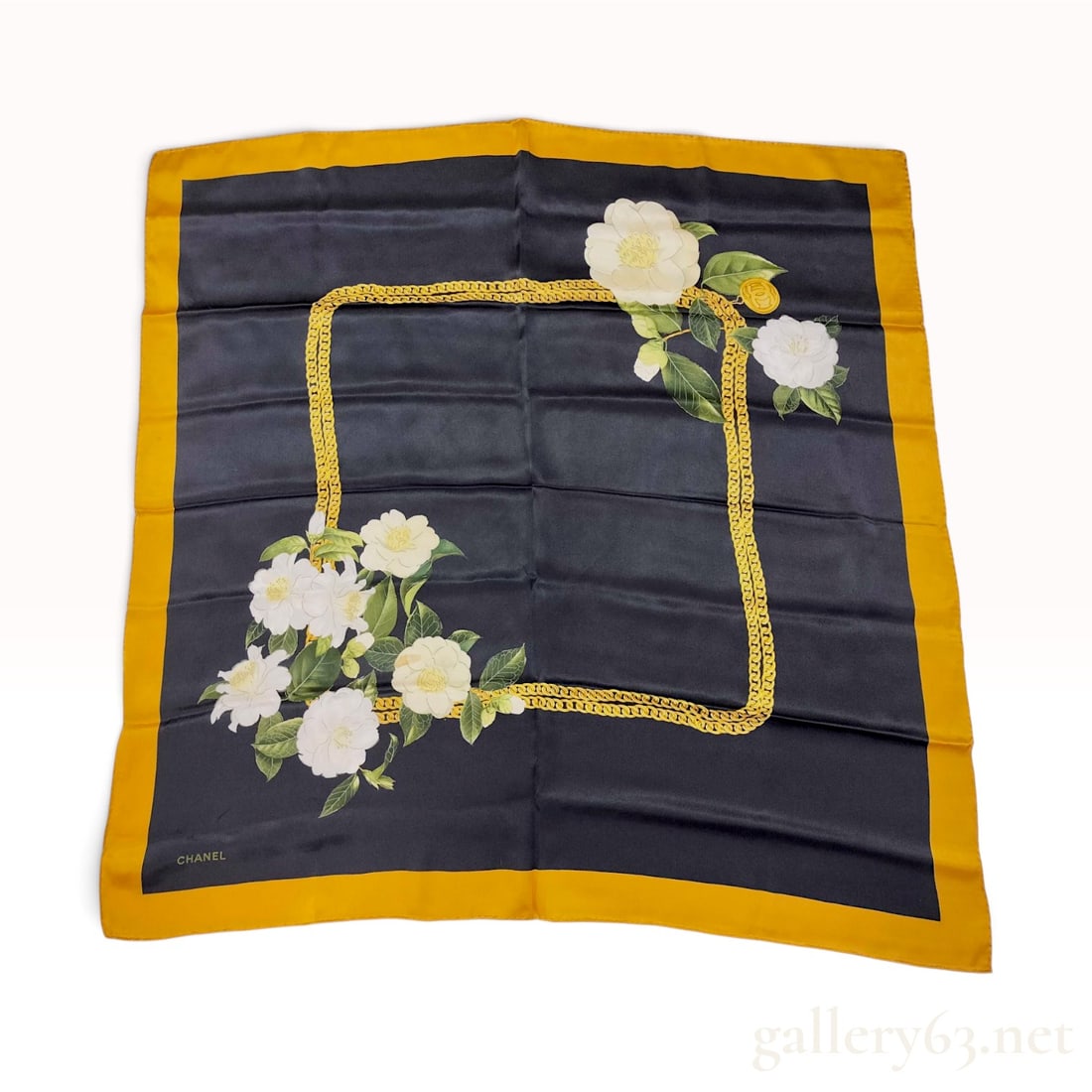 Chanel Camellia and Chain Print Silk Scarf: A Chanel square silk scarf featuring a black central field with a printed gold curb-link chain inner frame. The design is decorated with two clusters of white camellia flowers with green leaves. The u
