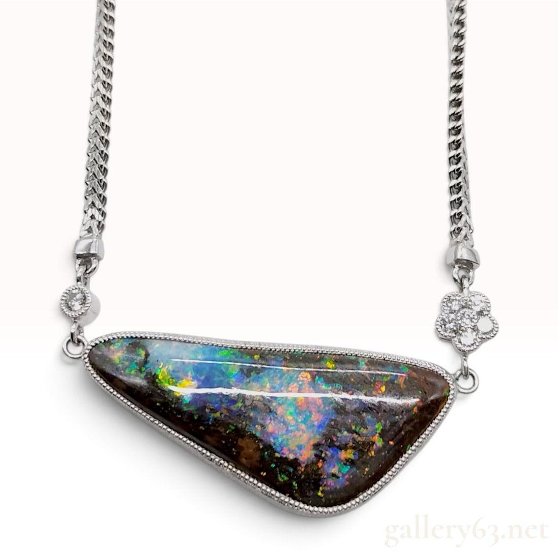 An Incredible 19.26ct Boulder Opal and Diamond Pendant Necklace: A necklace crafted from 18 karat white gold featuring a centered integral pendant with a large freeform triangular-cut boulder opal. The 19.26ct opal is bezel-set with a milgrain border and exhibits a