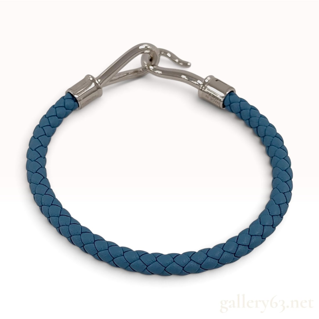 Hermes Blue Braided Leather Bracelet with Care Booklet: This Hermes bracelet is crafted from blue braided leather cord terminated with polished metal end caps. The piece utilizes a signature hook-and-eye closure mechanism. One metal end cap is impressed wi