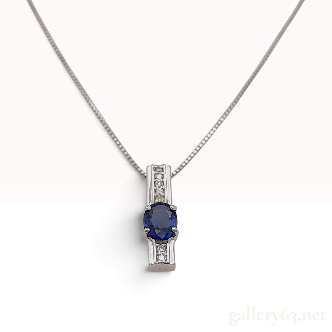 18 Karat White Gold Natural Blue Sapphire and Diamond Pendant Necklace: A contemporary 18 karat white gold necklace featuring a vertical bar pendant. The pendant centers one four-prong mounted oval-cut natural blue sapphire measuring approximately 6 x 4.5 mm. Flanking the