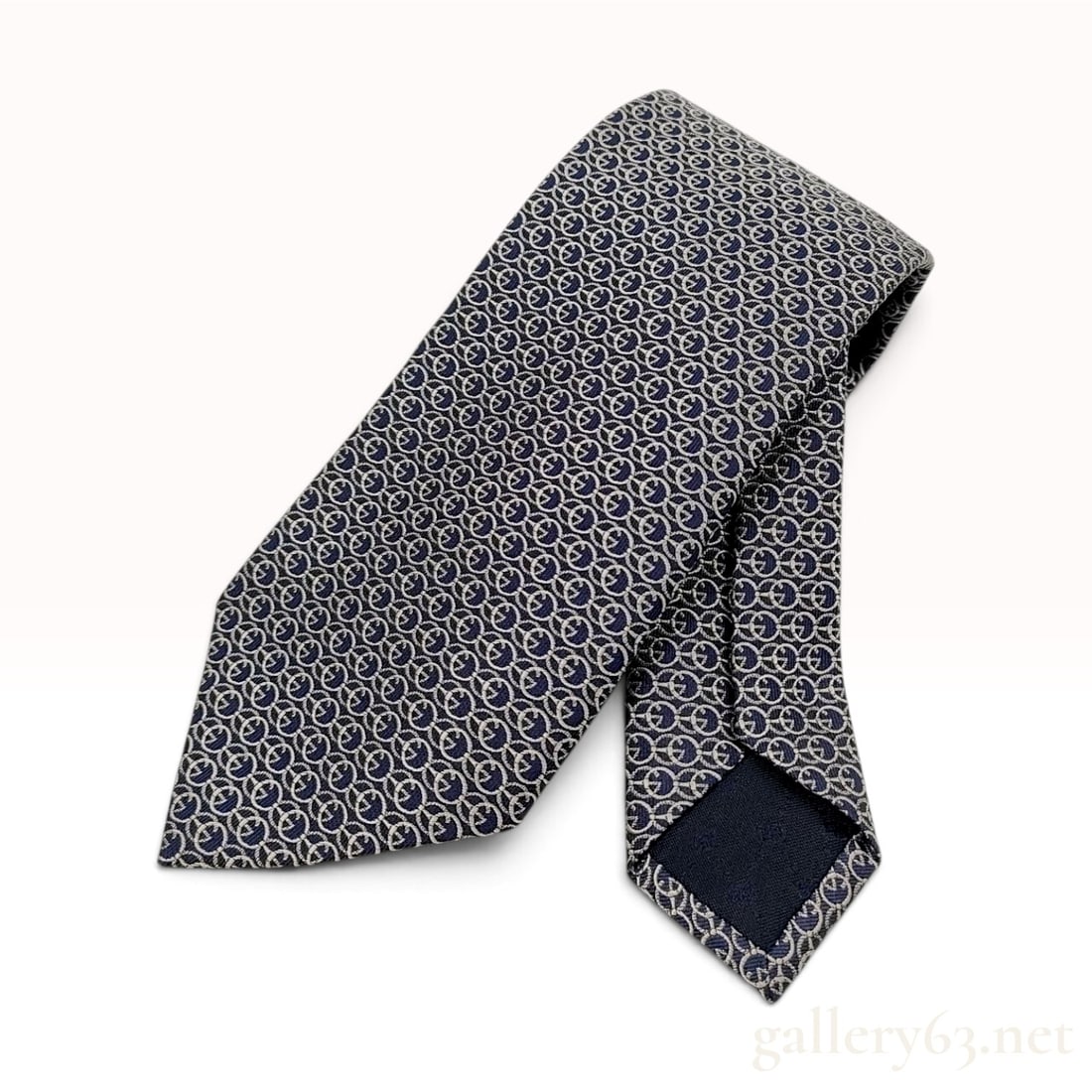 Gucci Interlocking G Chain-Link Navy Blue Silk Necktie: This Gucci necktie is crafted from 100% navy blue silk featuring a repeating silver-grey interlocking G monogram pattern arranged in a circular chain-link motif. The interior tip lining is finished in