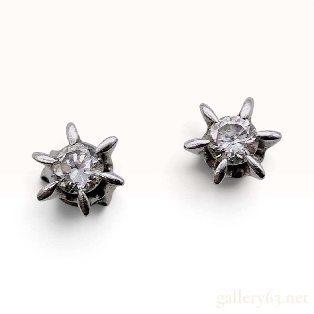 Platinum and Natural Diamond Starburst Stud Earrings: This is a matched pair of platinum stud earrings, each set with a round brilliant cut natural diamond. The gemstones are held within a stylized six-prong starburst setting, which features tapered, rou