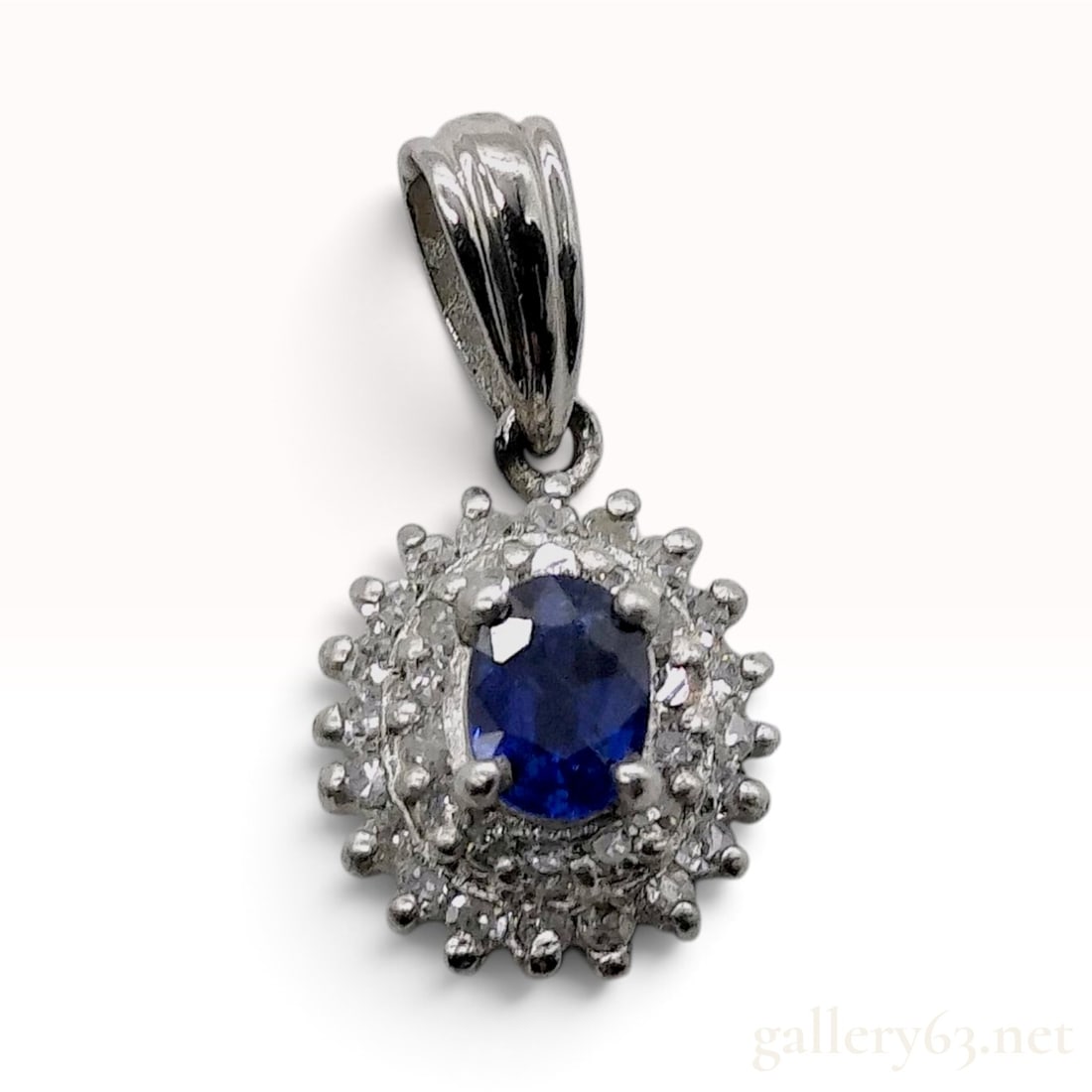 14 Karat White Gold 0.90ct Sapphire and 0.11ctw Natural Diamond Halo Pendant: This 14 karat white gold pendant features a central oval-cut natural blue sapphire secured in a four-prong setting. The primary stone is surrounded by a tiered sunburst-style double halo composed of a
