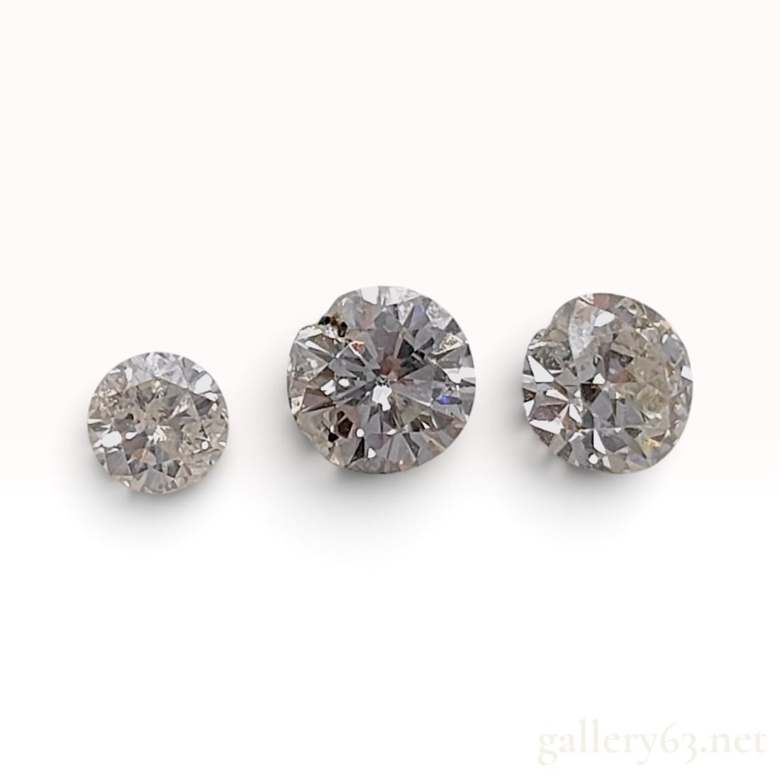 A Trio of Round Brilliant Cut Salt and Pepper Natural Diamonds: This trio of loose gemstones appears to be round brilliant-cut natural diamonds. The set features two larger stones of similar diameter and one smaller stone. Each diamond displays a salt and pepper c