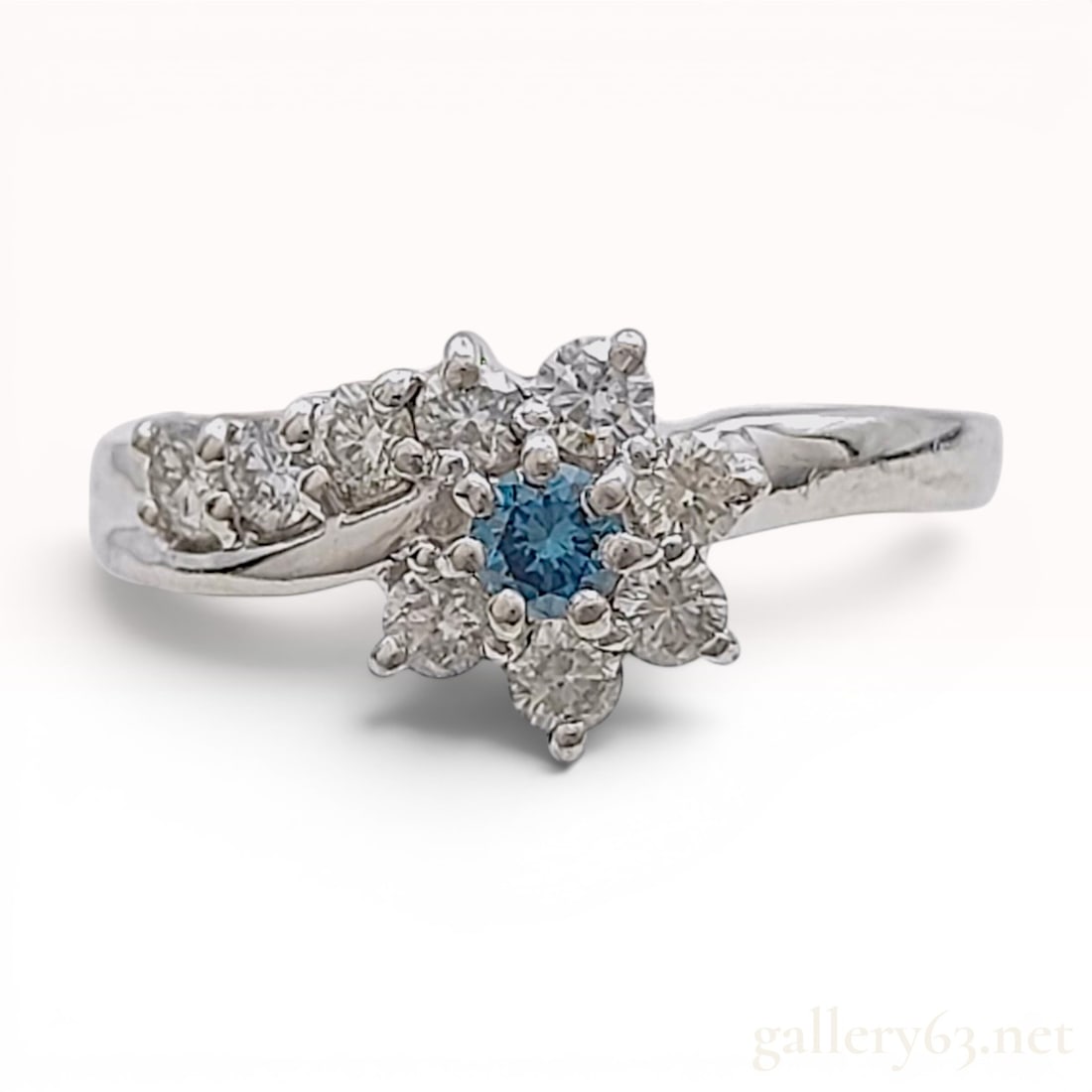 Platinum Flower Motif Bypass Ring with Irradiated Natural Blue Diamond and Diamond Accents: A platinum decorative ring featuring a floral cluster design. The center stone is a single round brilliant-cut natural diamond that has been artificially irradiated to a blue color. Surrounding the