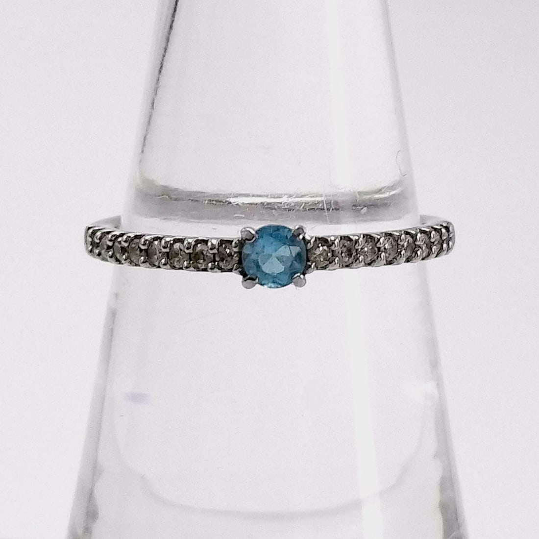 10K White Gold Ring with Diamonds and Round Cut London Blue Topaz: This ring is composed of 10-karat white gold and features a central round-cut light blue topaz held in a traditional four-prong setting. The shoulders of the shank are set with a series of small, roun