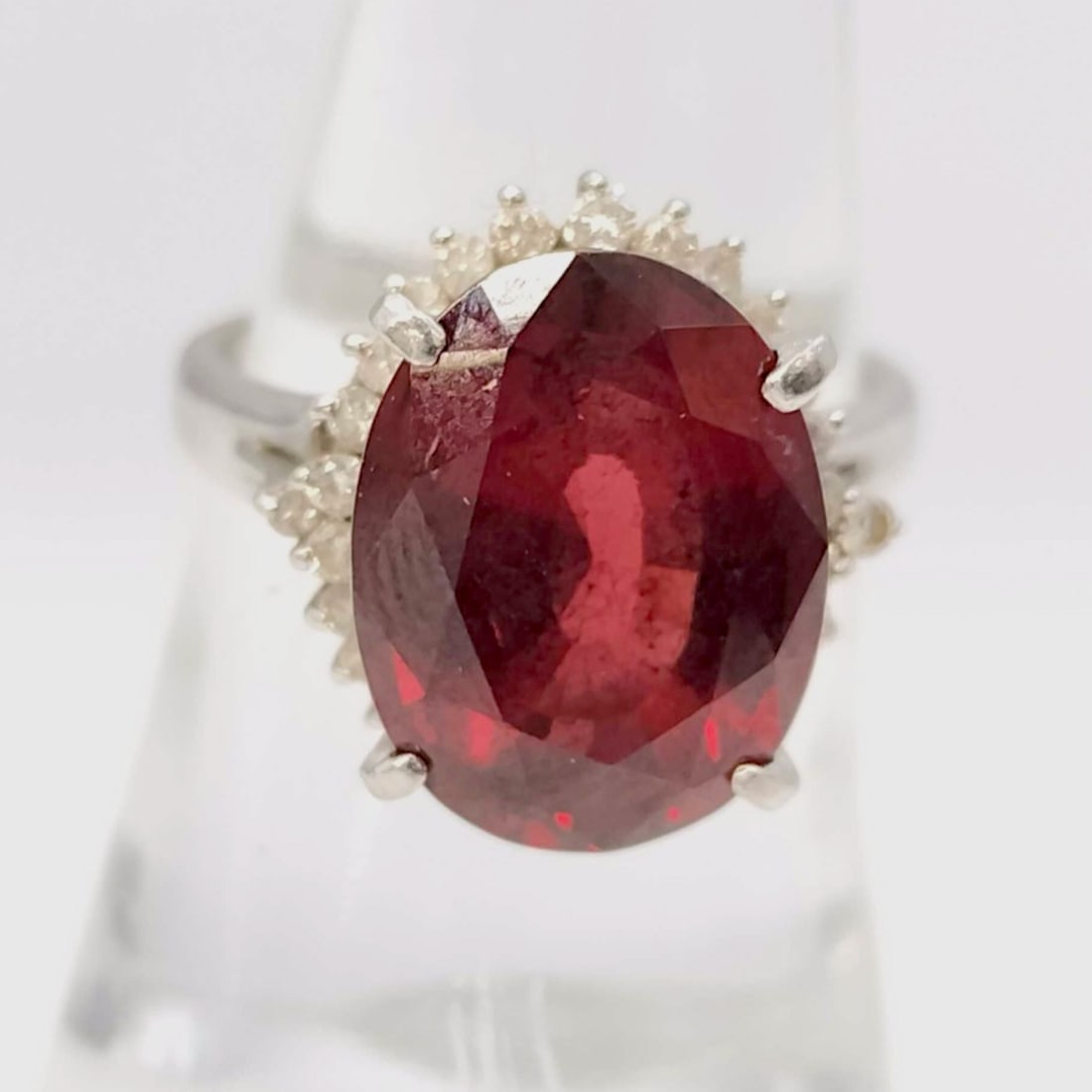 Platinum, Garnet and Diamond Halo Cocktail Ring: Platinum cocktail ring featuring a large oval-cut red gemstone secured in a double four-prong setting. The center stone is surrounded by a halo of round brilliant-cut diamonds atop an ornate openwork 