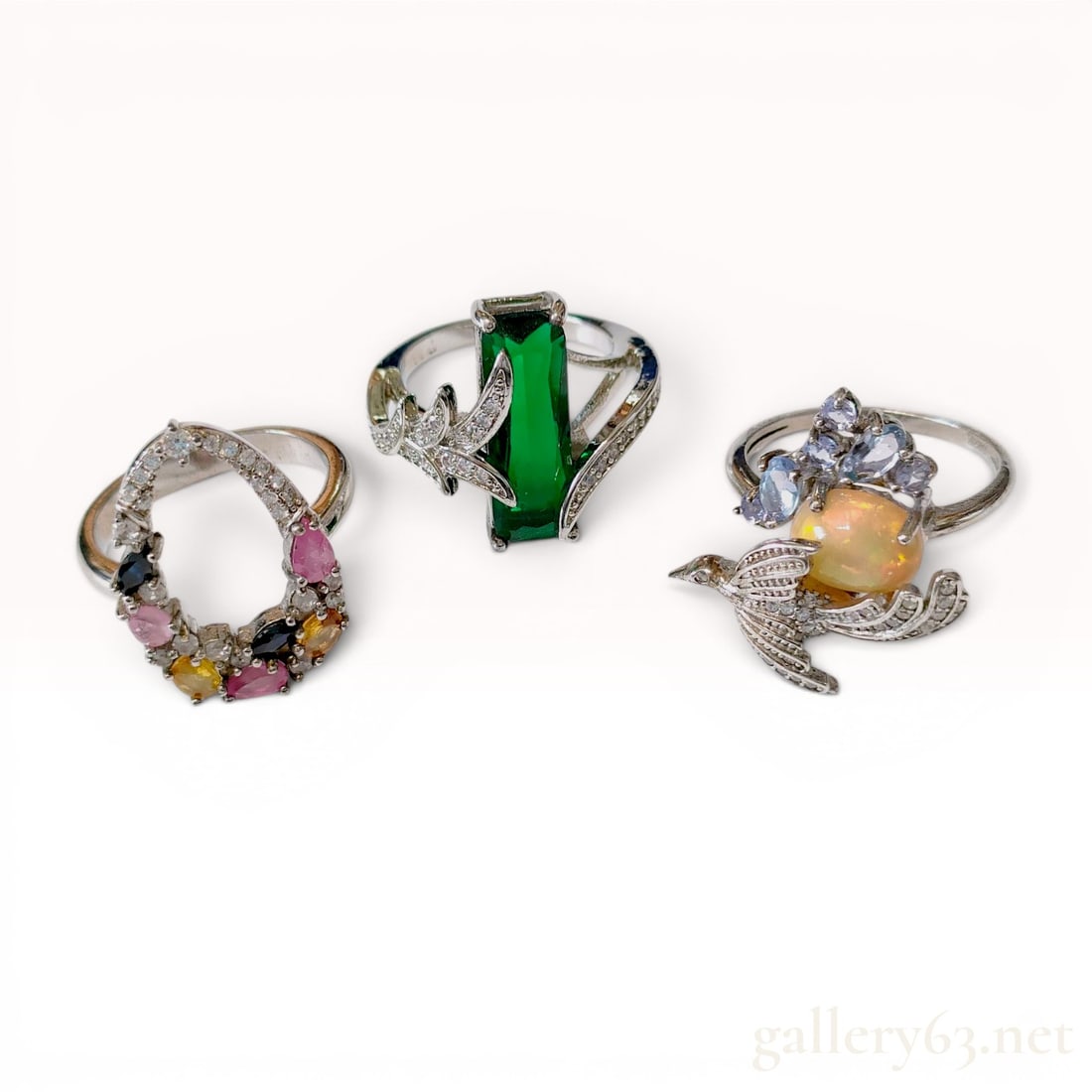Three Sterling Silver and Multi-Gemstone Cocktail Rings: A collection of three sterling silver cocktail rings featuring various gemstone motifs including a hummingbird, a large emerald-cut green stone, and a multi-colored open teardrop design. The rings are