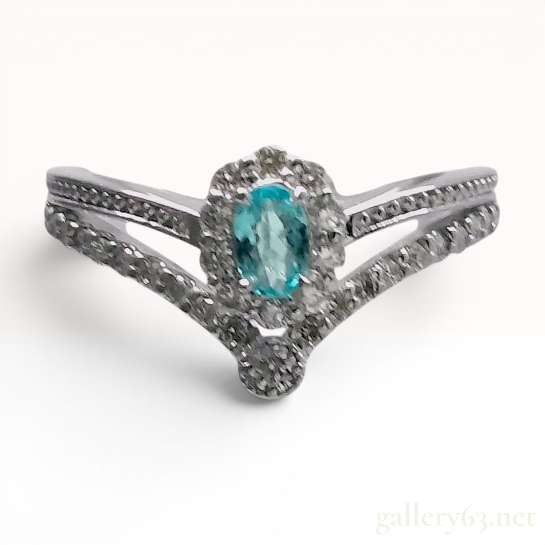 Platinum Paraiba Tourmaline and Diamond Halo Chevron Ring: A platinum fashion ring featuring a central oval fancy-cut greenish-blue tourmaline weighing 0.125 carats. The gemstone is set within a diamond-encrusted halo. The ring is constructed with a split-sha