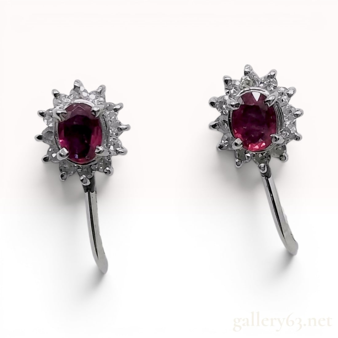 A Pair of Platinum, Ruby, and Diamond Cluster Screw-Back Earrings: A matched pair of ruby and diamond cluster earrings featuring a non-pierced screw-back design. Each earring centers an oval-cut ruby of a deep pinkish-red hue with visible natural inclusions. The rubi