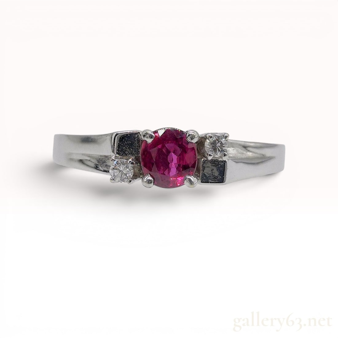 14kt White Gold Ruby and Diamond Dress Ring: 14 karat white gold multi-stone dress ring featuring a central round-cut ruby and two offset round-cut diamond accents. The gemstones are secured in prong settings atop split shoulders that integrate 
