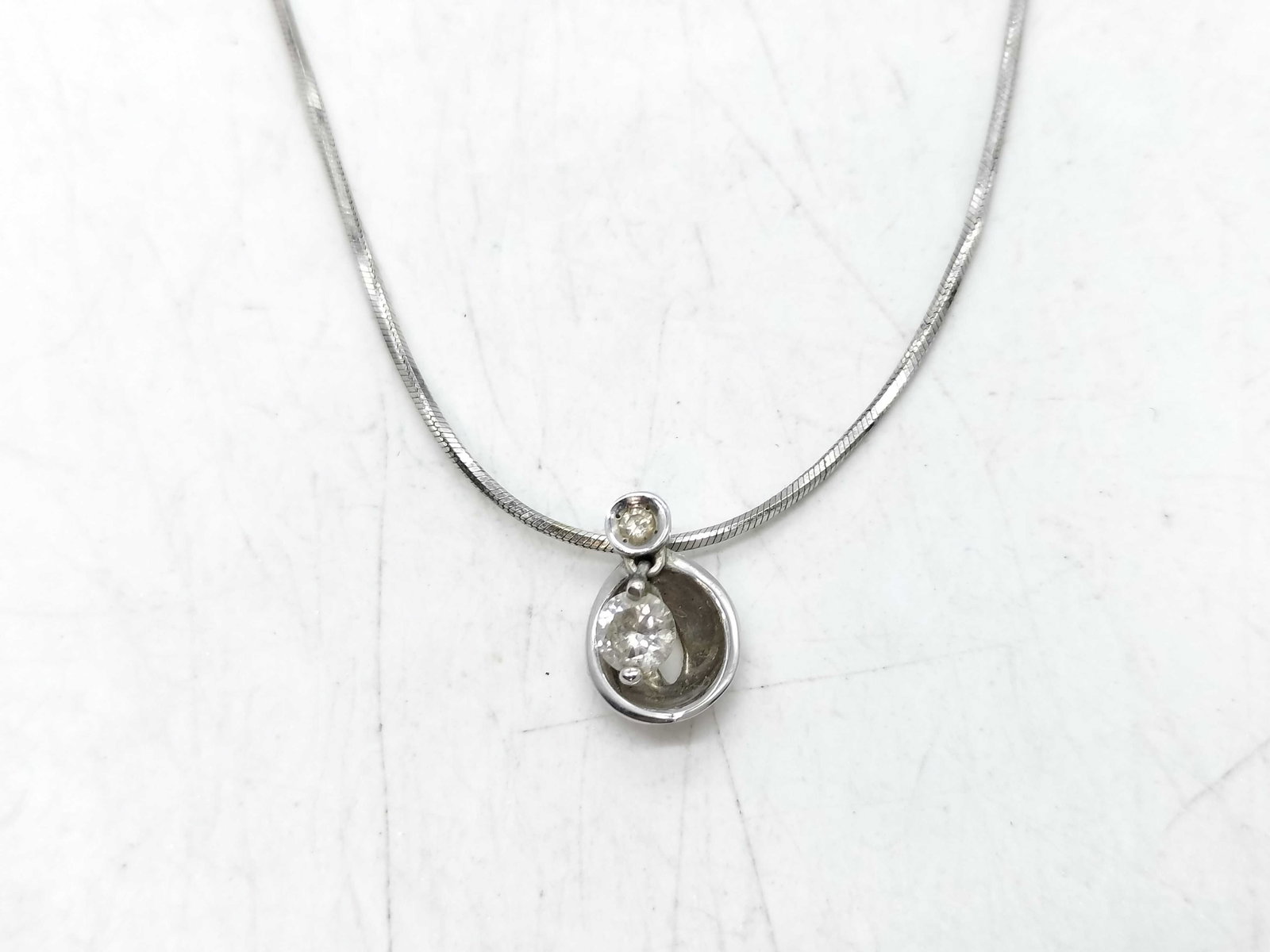 10K White Gold and Diamond Pendant Necklace: A minimalist drop pendant necklace crafted in 10-karat white gold. The pendant features a two-part design, with a small bezel-set round diamond at the top suspending a larger, cradle-style open settin