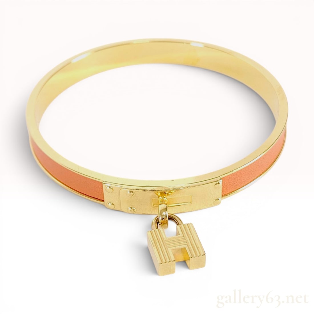 Hermes Kelly H Cadena Bangle with Orange Leather Inlay and Original Box: This Hermes Kelly H Cadena bangle bracelet is constructed of gold-toned metal with a signature orange leather inlay. The design features a central hardware plate modeled after the Kelly bag closure, f
