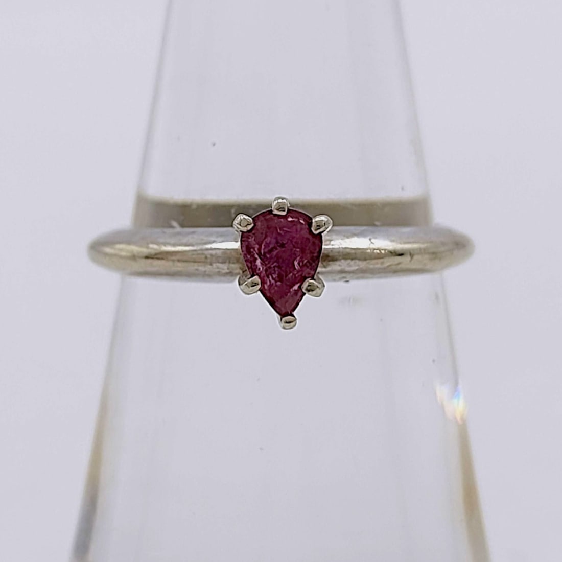14 Karat White Gold Pear-Cut Ruby Solitaire Ring: 14 karat white gold solitaire ring featuring a pear-cut reddish-pink ruby. The stone is secured in a five-prong setting on a simple polished band. The interior of the shank is stamped with a