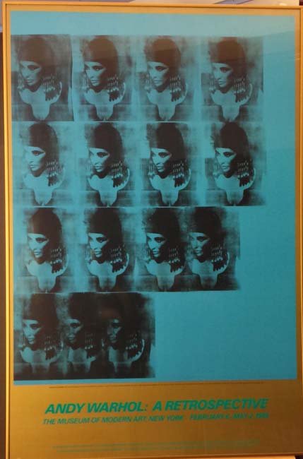 Elizabeth Taylor Cleopatra Exhibition Poster after Andy: This rare, framed exhibition poster features an image, many images actually, of Elizabeth Taylor dressed as Cleopatra. The black images are printed over a flat, blue plane of color which is framed by 