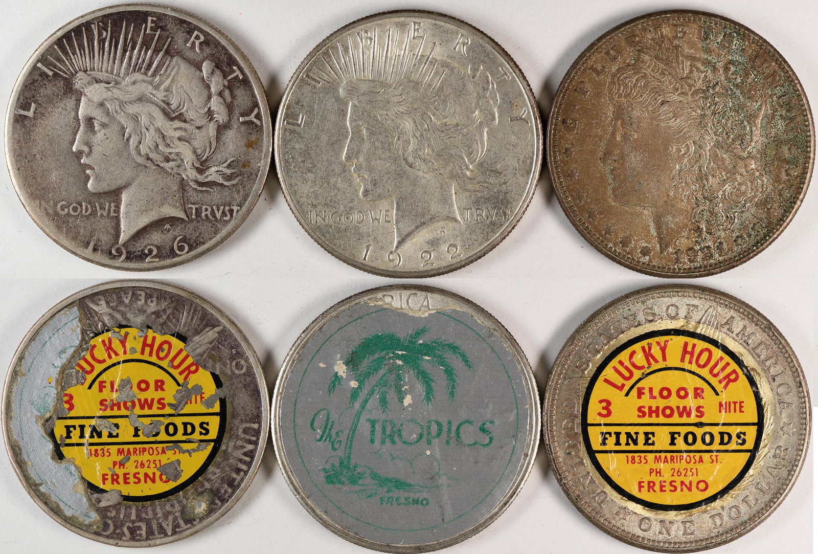 Sticker Silver Dollars Group (120275): Three Lady Liberty silver dollars with stickers: Lucky Hour/Floor Shows/3 Nite/Fine Foods/1835 Mariposa St/Ph. 26251/Fresno, c1921; The Tropics/(pictorial palm tree)/Fresno, c1922; Lucky Hour/Floor Sh