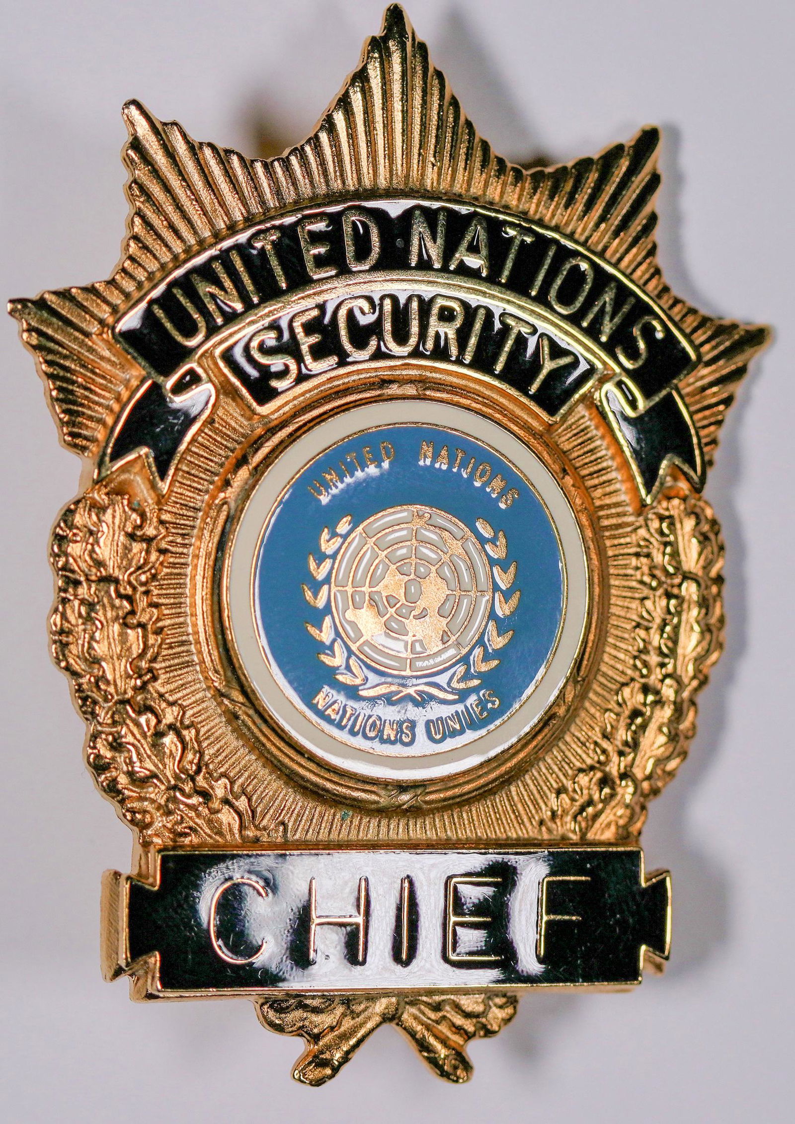 United Nations Security Chief Badge [132480]: This is a United Nations Security Chief badge in gold tone, sunburst design. The UN logo is present in a robin's egg blue background. About 3 in. long. It has a strong pin back clasp with no hallmark.