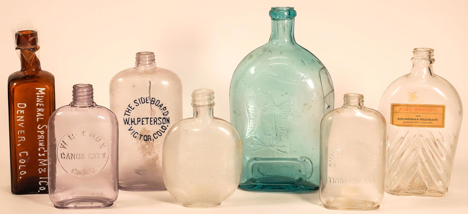 Variety of Colorado Whiskey Flasks from a Variety of Locations [162618]: Lot of seven. 1) The Side Board / WH Peterson / Victor, Colo. Pint pocket flask. Cracked face. 2) Half pint. Simon Sanders & Co. / Trinidad. Crack at base of neck. 3) For Pikes Peak / with hobo. Quart