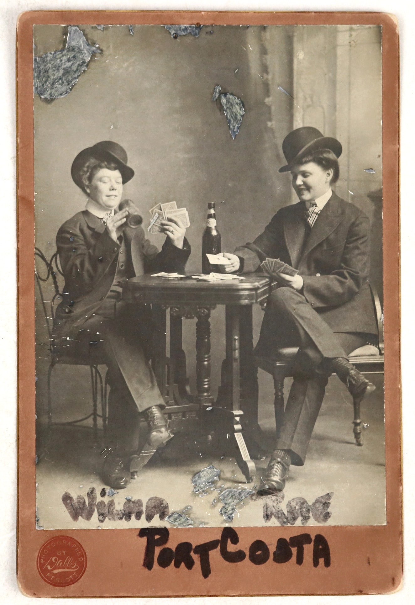 Gambling Women, Vintage Photograph [195055]: A posed photo of two women in suits and bowler hats drinking and playing cards. The women are labelled 'Wilma' and 'Rae' with the name of the town where the photo was taken, Port Costa, written below.