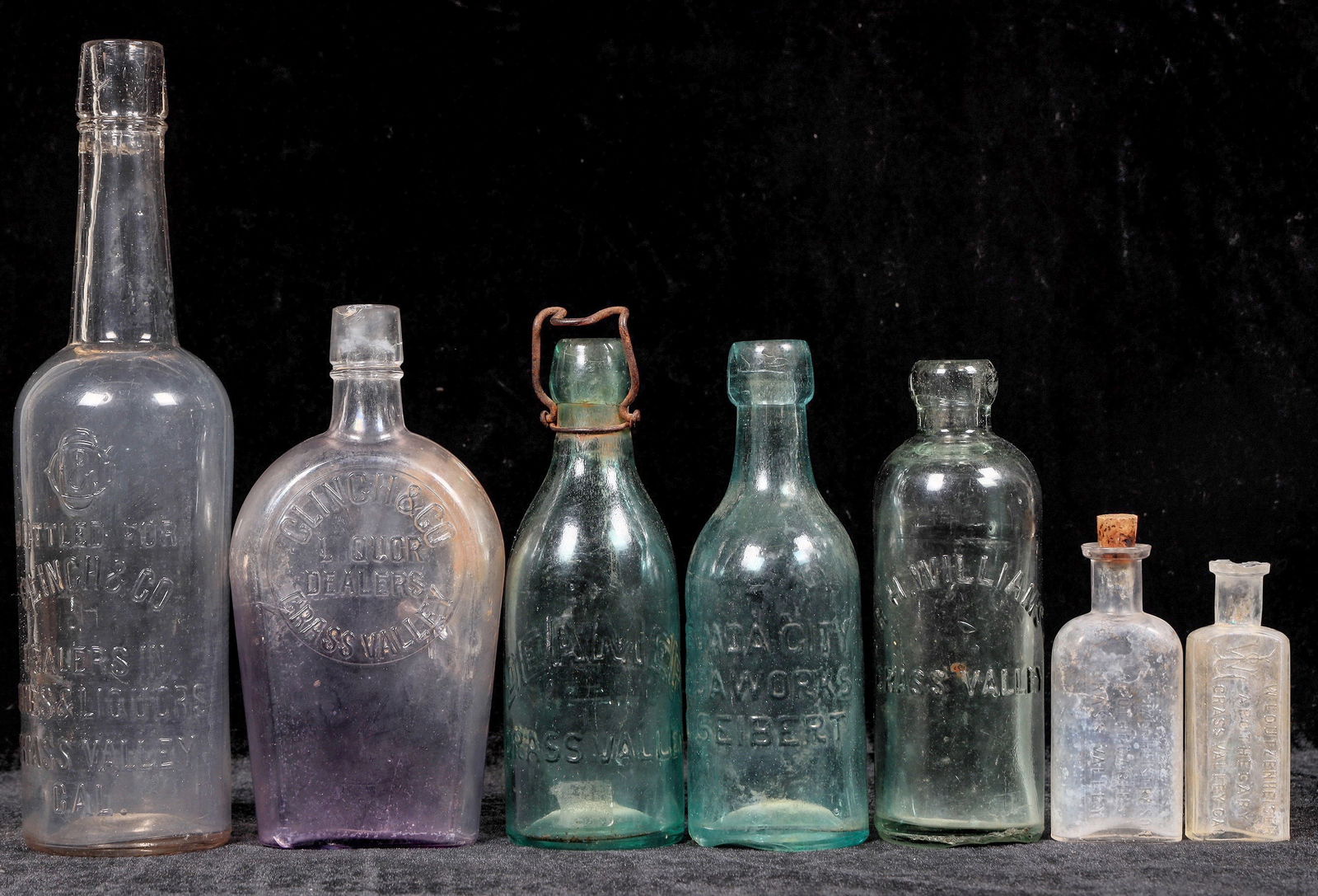 Grass Valley Bottle Group w/ Problems [194398]: Group of 7 Grass Valley embossed bottles: 1. Clinch, clear sixth, neck wear 2. Clinch, quart flask, cracks 3. Siebert, blob soda, crack 4. Deamer, blob soda, lip chip 5. Williams, Hutch, base chip 6 &