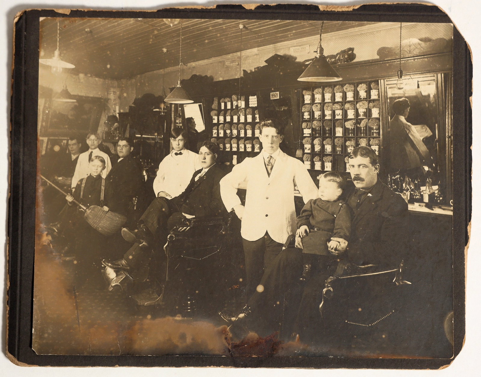 Shaving Mug/Barber Interior [200415]: Barber shop interior with about 50 personalized shaving mugs in the back, names not in focus. Location unknown, though self ID claims Goldfield as the location. The fourth man from the left bears a st