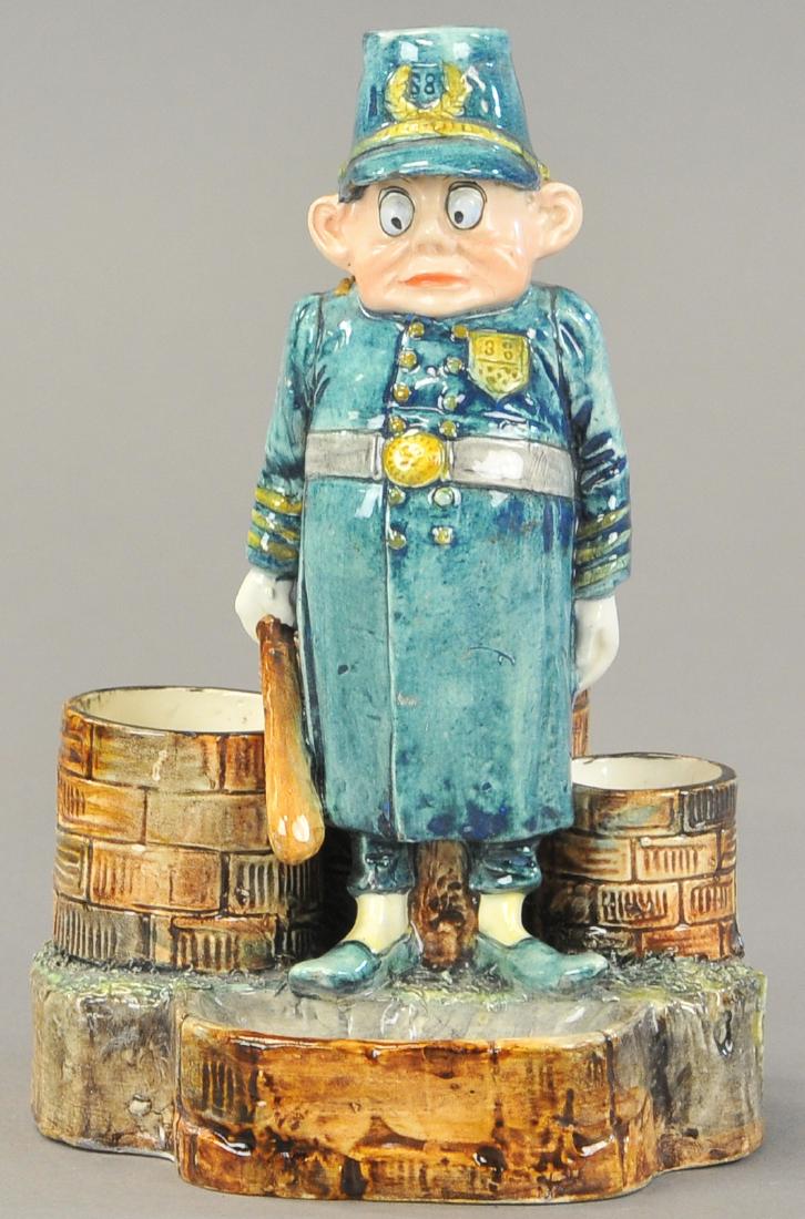 MAJOLICA BROWNIE MATCH HOLDER: 104. MAJOLICA BROWNIE MATCH HOLDER | A Palmer Cox English Bobby w/cigar, cigarettes, match holders and a candle holder cap on his head. 9" h.