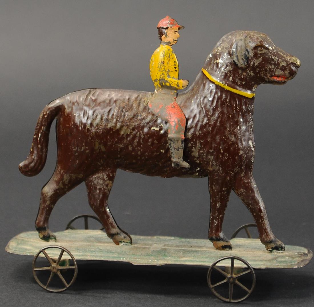 JOCKEY RIDING LARGE DOG: 183. JOCKEY RIDING LARGE DOG | Attributed to Althof Bergmann c. 1880s, familiar elegant form of large hand painted tin dog on green platform, with man rider on back, spoke wheels, this pull toy was ve